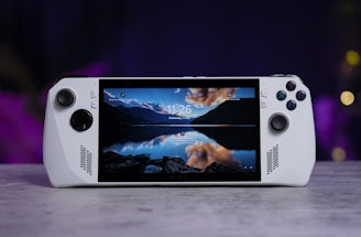 A handheld gaming console with a central screen displaying a scenic wallpaper of mountains and a lake, flanked by controller buttons and joysticks on both sides. The lighting around is dim, with purplish hues in the background, creating a modern and elegant atmosphere.